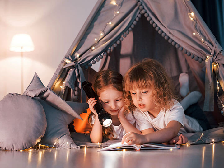Two young girls laying down in a homemade tent with decorative string lights reading a book with a flashlight