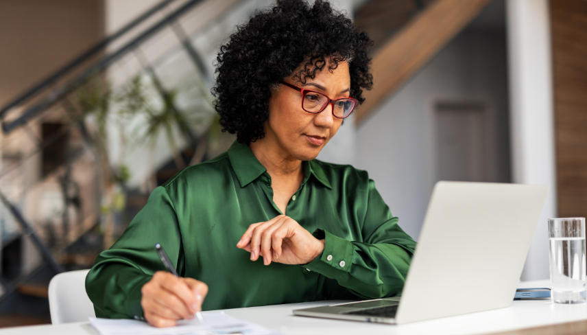 A woman in glasses writing notes while working on a laptop at a desk.